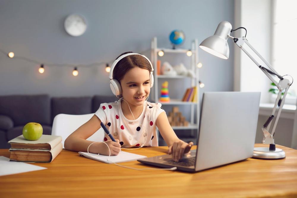 A picture of a girl in her bedroom wearing a white shirt with polka-dots attending a virtual tutoring session on a silver laptop.